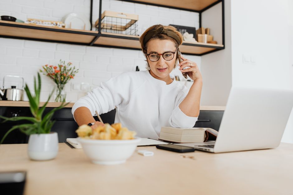 Femme au téléphone dans une cuisine, prenant des notes d'une recette tout en consultant un livre de cuisine.