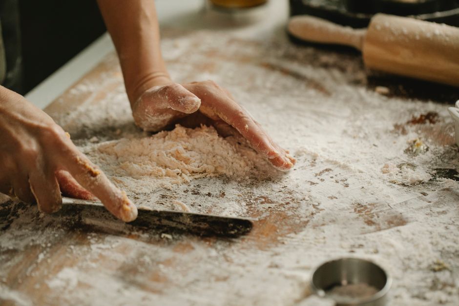 Mains pétrissant de la pâte sur un plan de travail recouvert de farine. Ustensiles de cuisine en arrière-plan.