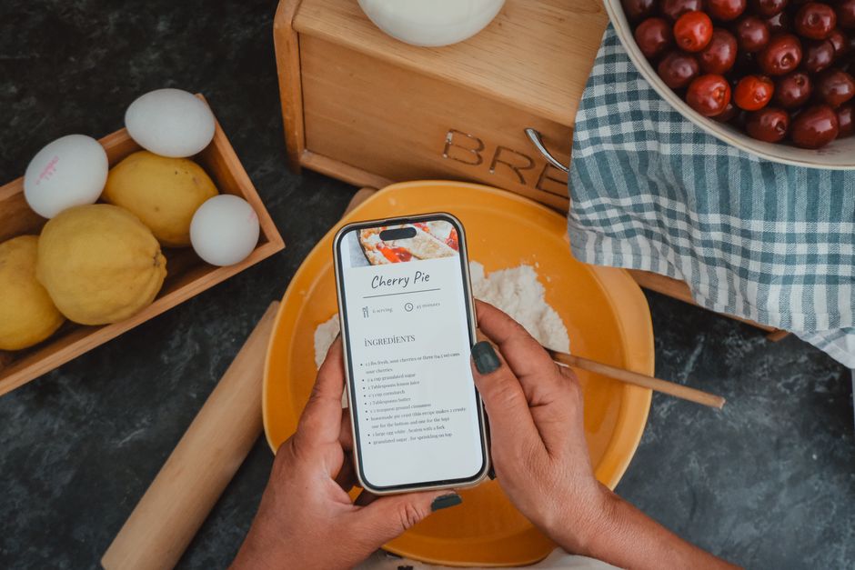 Un smartphone affichant une recette de tarte aux cerises, posé sur une table avec des ingrédients comme des œufs, des citrons